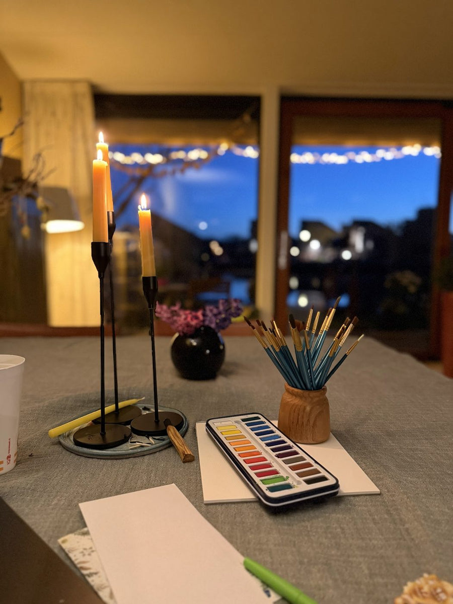 Candles, a watercolour set and paper on a table at dusk