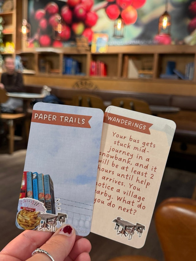 A hand holding a Paper Trails Wanderings card in a cozy cafe